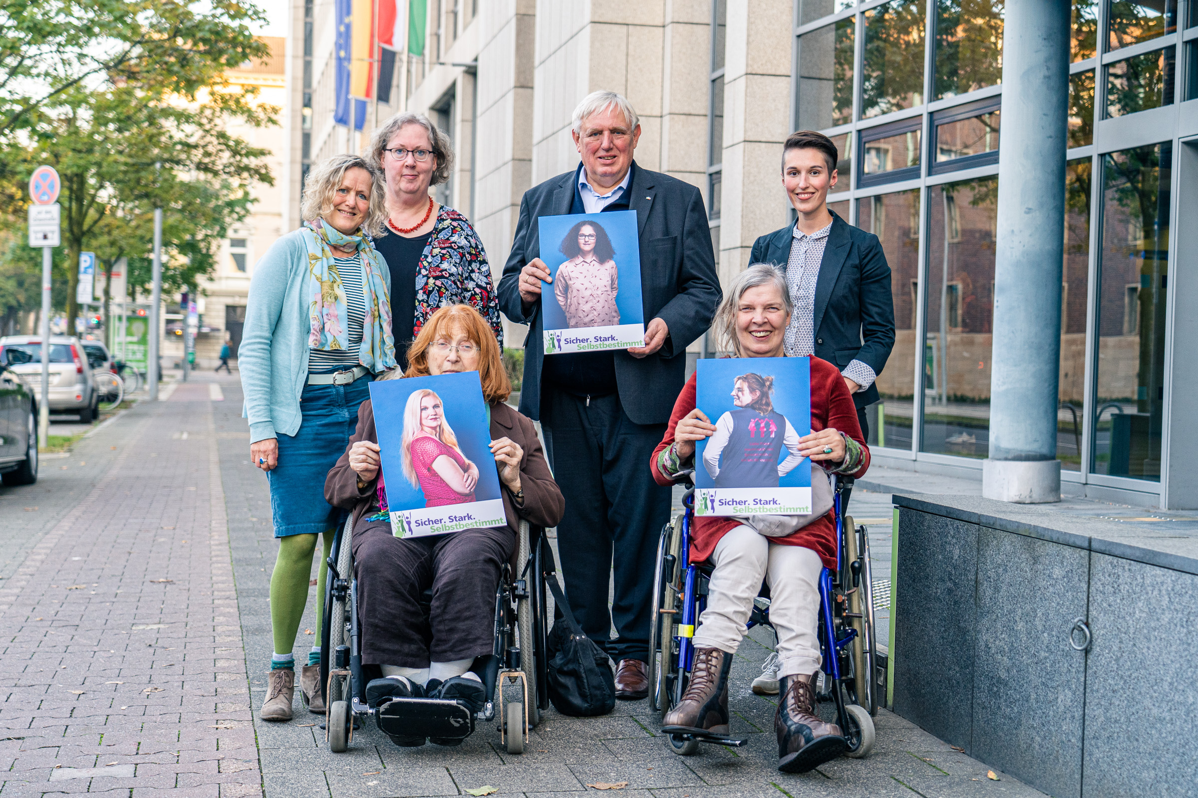 Die fünf Vertreter*innen des Netzwerks und Netzwerkbüros Frauen und Mädchen mit Behinderung/chronischer Erkankung stehen zusammen mit Minister Laumann vor dem Ministeriumsgebäude. Zwei Vertreterinnen sind im Rollstuhl, die anderen stehen dahinter. Minister Laumann steht in der Mitte und hält ein Plakat in die Kamera. Alle blicken freundlich in die Kamera.